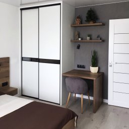 OLDI DESIGN SPÓŁKA Z OGRANICZONĄ ODPOWIEDZIALNOŚCIĄ - Nowoczesna sypialnia z łóżkiem, szafą przesuwną z białymi frontami i czarnymi ramami, drewnianym biurkiem z szarą welurową krzesłem oraz półkami na ścianie w kolorze szarym.
