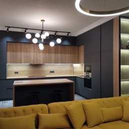OLDI DESIGN SPÓŁKA Z OGRANICZONĄ ODPOWIEDZIALNOŚCIĄ - Nowoczesna kuchnia z ciemnoszarymi szafkami, drewnianymi frontami, wyspą z hokerami i oświetleniem punktowym, widziana z perspektywy salonu z żółtą sofą.