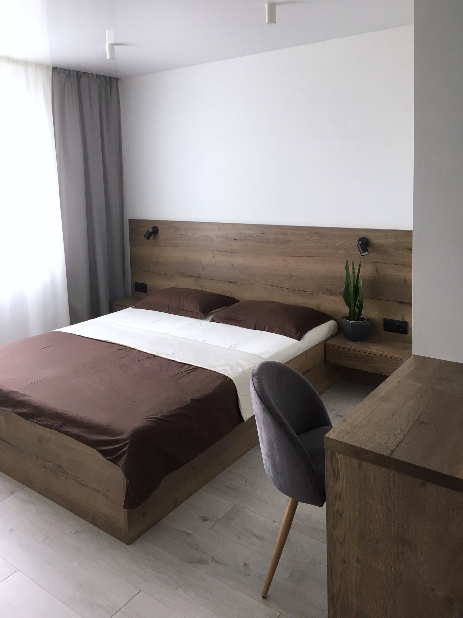 Nowoczesna sypialnia z drewnianym łóżkiem, szafką nocną z lampką i rośliną, szarą zasłoną i krzesłem na drewnianej podłodze.