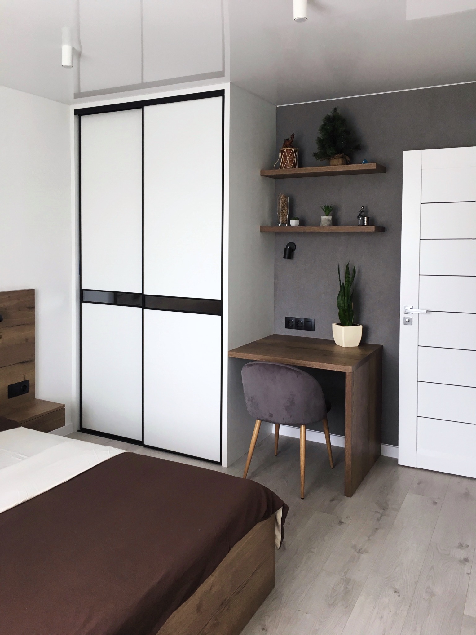 Nowoczesna sypialnia z łóżkiem, szafą przesuwną z białymi frontami i czarnymi ramami, drewnianym biurkiem z szarą welurową krzesłem oraz półkami na ścianie w kolorze szarym.