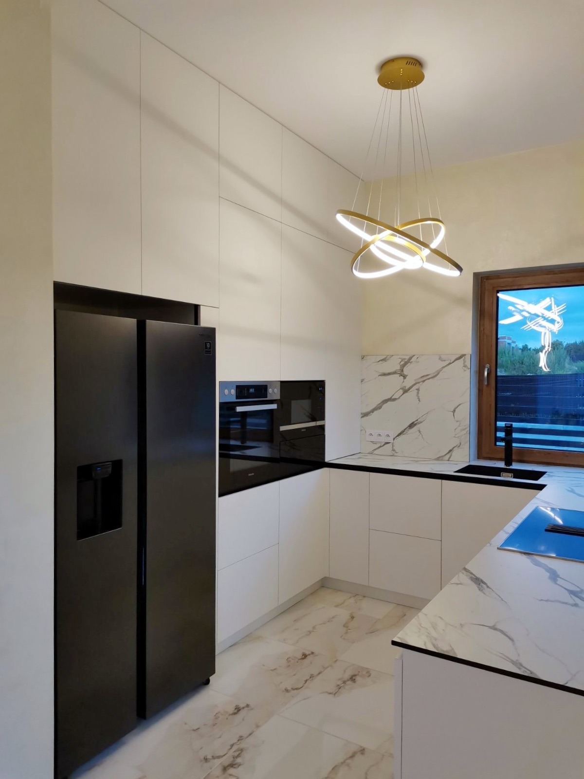 Nowoczesna kuchnia z białymi, gładkimi frontami szafek, czarnym sprzętem AGD (lodówka side-by-side i piekarnik), blatem imitującym marmur, złoty żyrandol z okręgów i oknem z widokiem na zewnątrz.