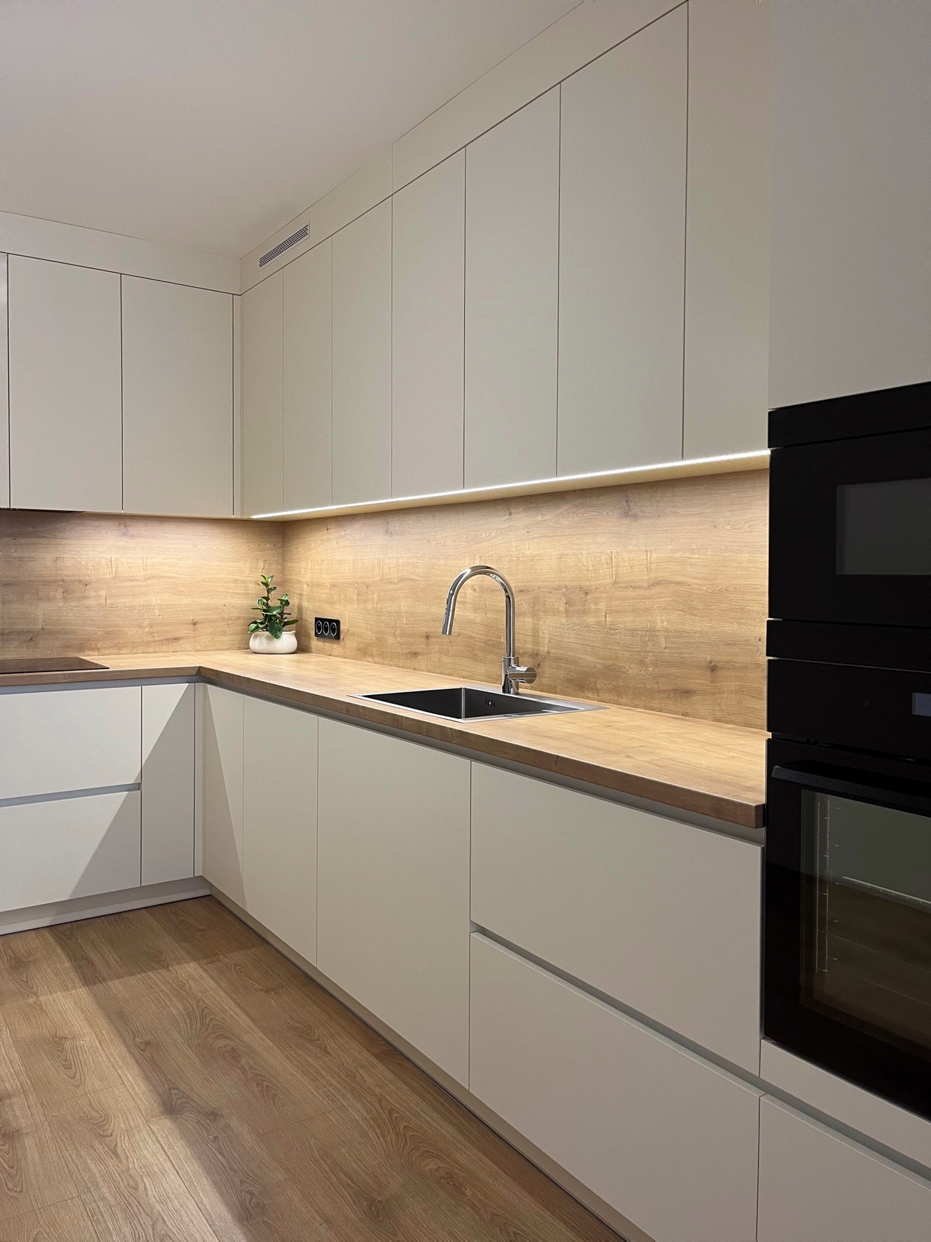 Nowoczesna, minimalistyczna kuchnia z jasnymi, gładkimi frontami szafek, drewnianym blatem i oświetleniem LED pod szafkami wiszącymi, z czarnym piekarnikiem w zabudowie i małą roślinką w doniczce...