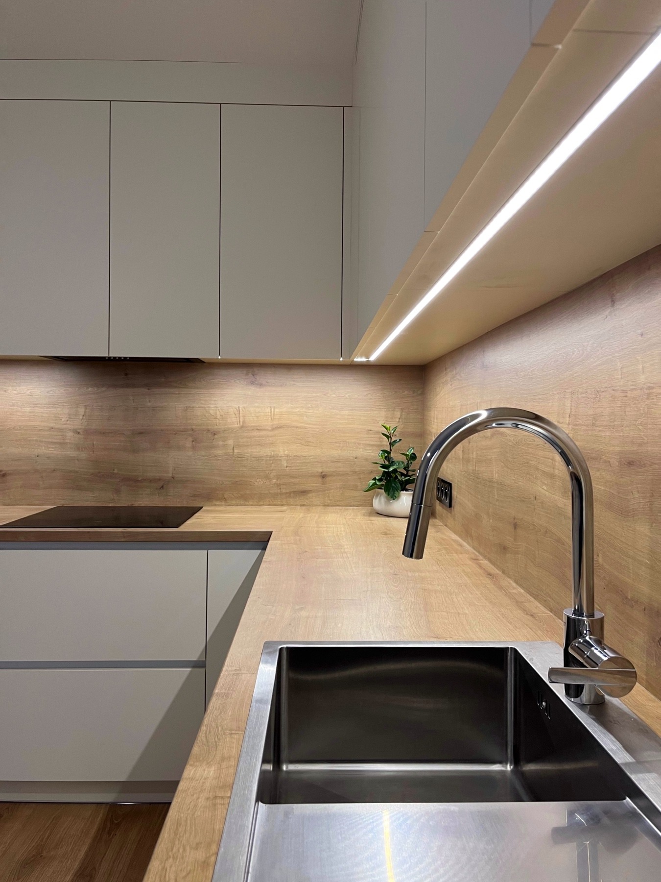 Nowoczesna kuchnia z jasnymi, gładkimi frontami szafek, drewnianym blatem i panelem ściennym, podświetleniem LED oraz stalowym zlewem i baterią.