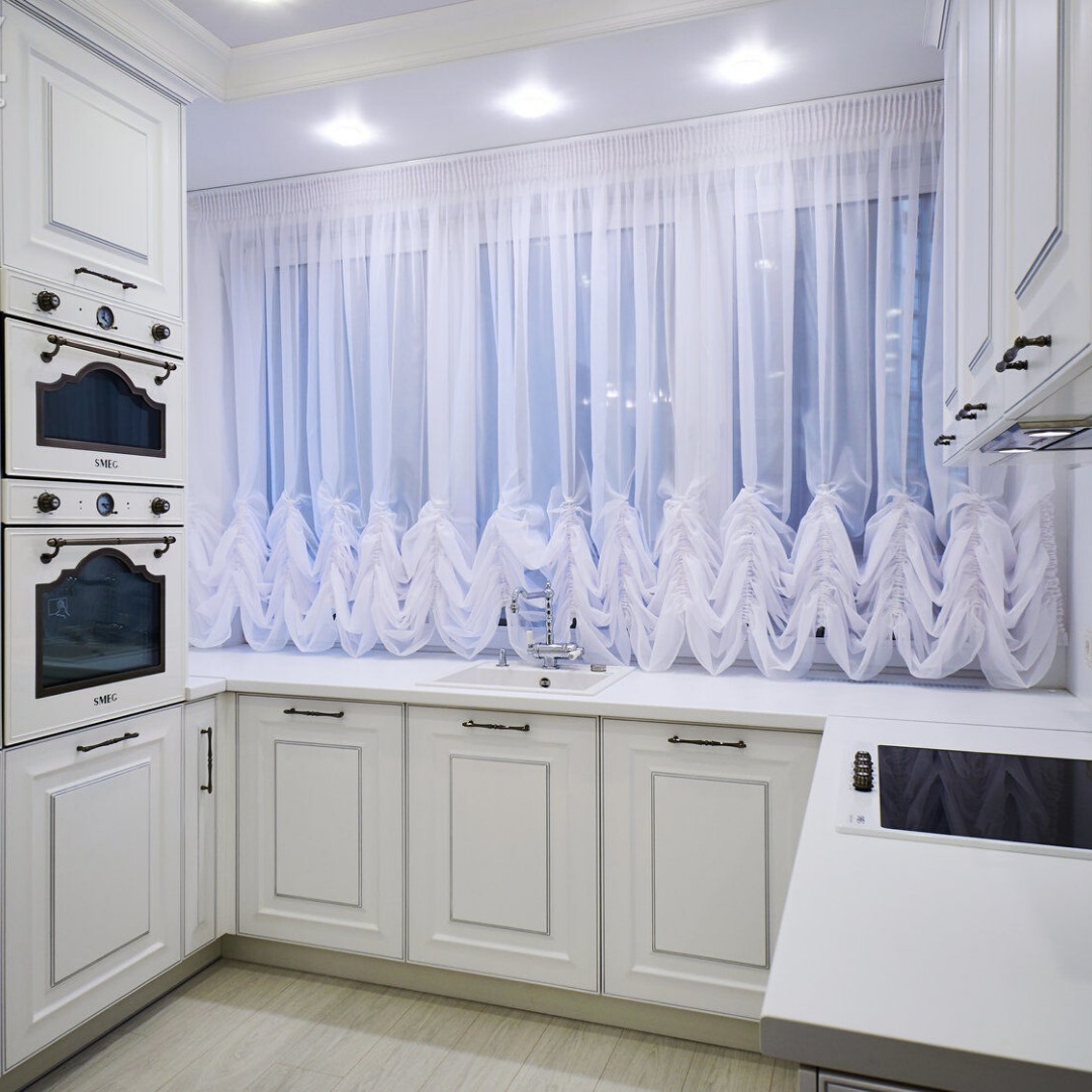 Elegancka, biała kuchnia z klasycznymi frezowanymi frontami, wyposażona w zabudowane piekarniki marki SMEG i zwiewne, dekoracyjne firanki w oknie nad zlewem.