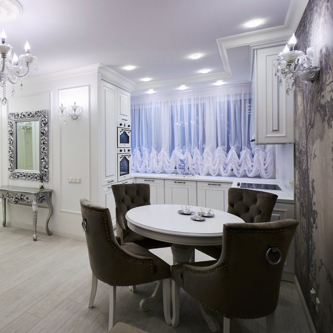 Elegancka, jasna kuchnia w stylu glamour z białymi szafkami, okrągłym stołem z brązowymi krzesłami i dekoracyjnym lustrem w srebrnej ramie.