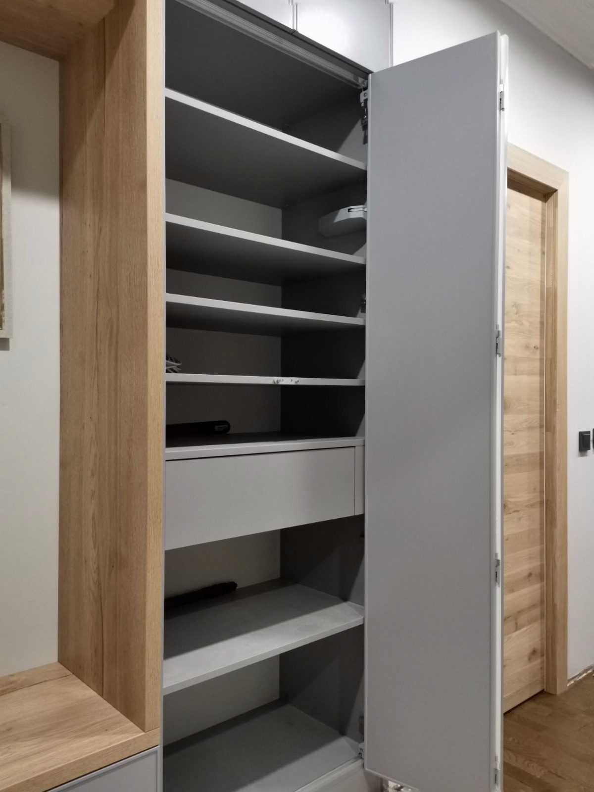 Otwarta szafa wnękowa z szarymi półkami i szufladą, obudowa z drewna w odcieniach beżu, widoczny mechanizm otwierania drzwi, drewniana ościeżnica drzwi w tle.