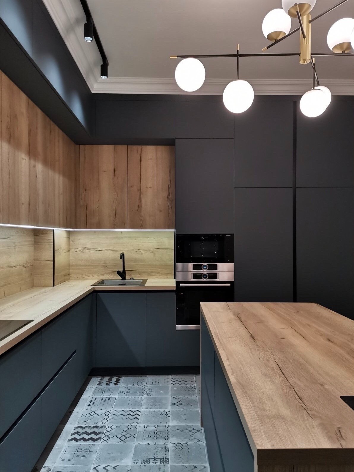 Nowoczesna kuchnia z szarymi szafkami i drewnianymi blatami, oświetlona designerską lampą sufitową z kulistymi kloszami, z wzorzystą podłogą.