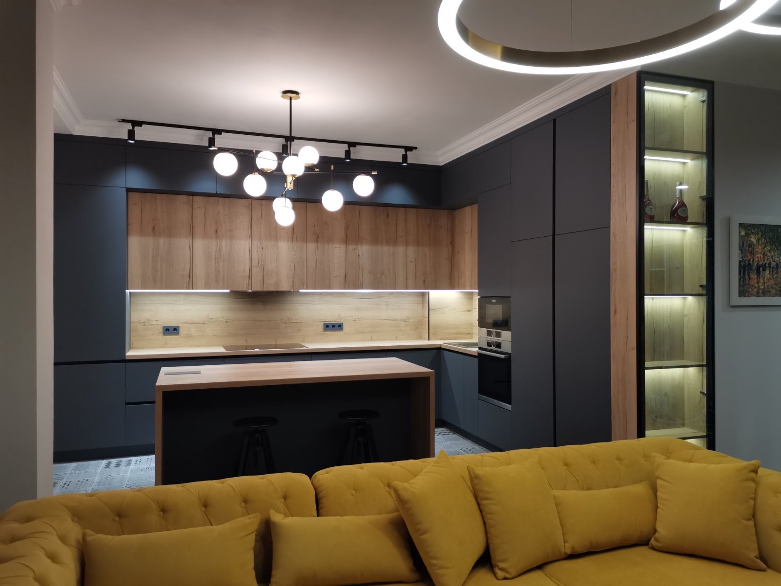 Nowoczesna kuchnia z ciemnoszarymi szafkami, drewnianymi frontami, wyspą z hokerami i oświetleniem punktowym, widziana z perspektywy salonu z żółtą sofą.