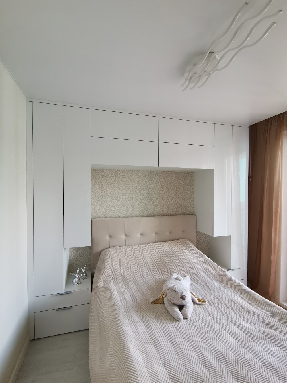 Nowoczesna sypialnia z białymi szafami w zabudowie nad łóżkiem, beżowym zagłówkiem i pluszowym misiem na narzucie w jodełkę.