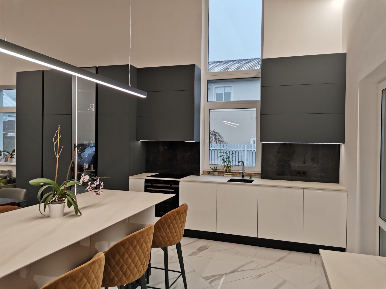 Nowoczesna kuchnia z czarnymi szafkami górnymi i białymi dolnymi, wyspą z blatem z jasnego kamienia, oświetleniem LED i marmurową podłogą.