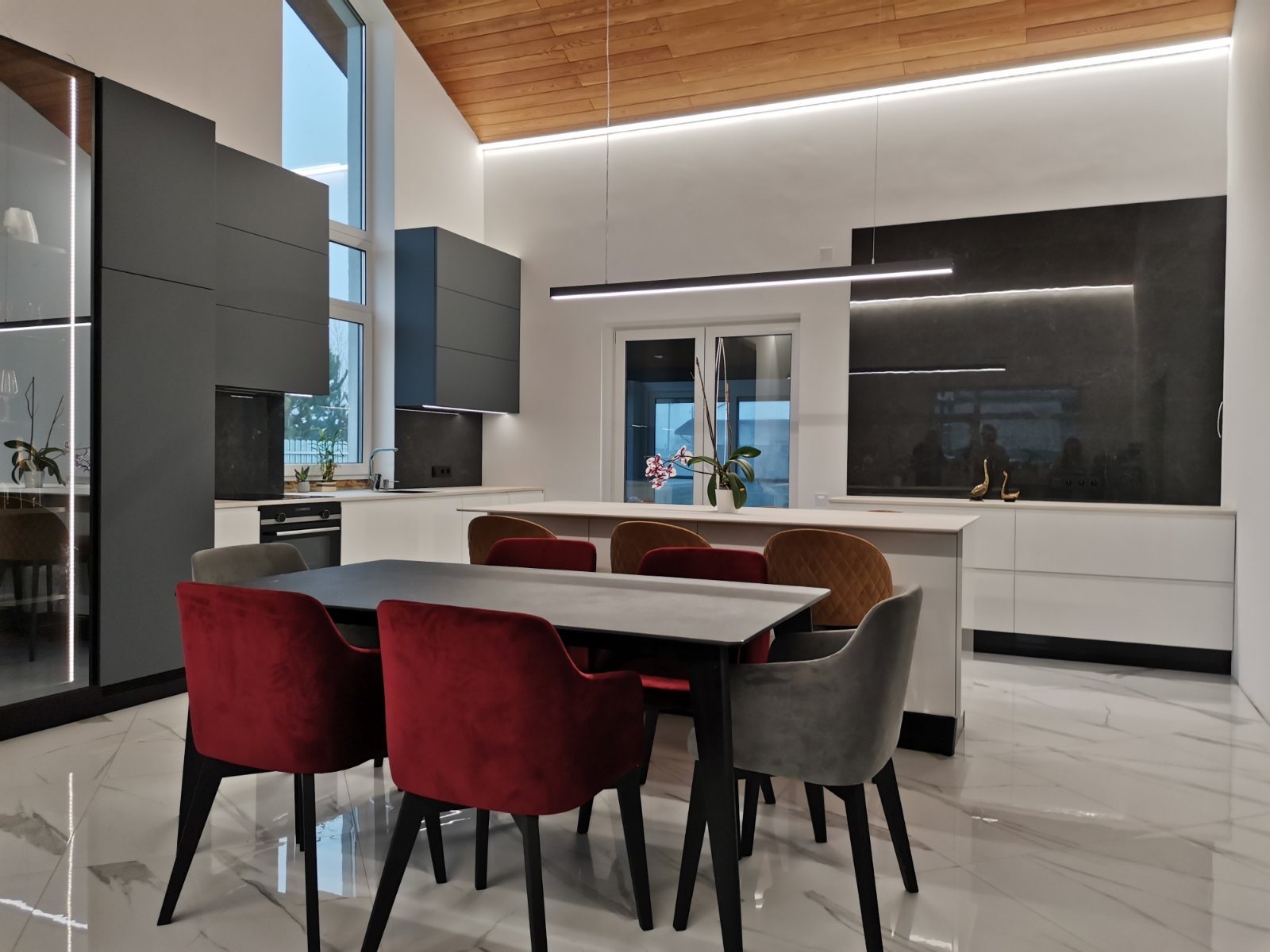 Nowoczesna kuchnia z szarymi szafkami, białym blatem i wyspą, oświetleniem LED, marmurową podłogą i stołem z krzesłami w kolorze czerwonym i szarym