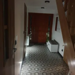 Korytarz z wzorzystą, czarno-białą podłogą, drewnianymi drzwiami wejściowymi i schodami, białą ławką z szarą poduszką oraz dekoracjami w postaci roślin w doniczce i wieszaka na ubrania.