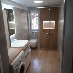 Nowoczesna łazienka z drewnopodobnymi płytkami, kabiną prysznicową typu walk-in, umywalką nablatową z pralką pod spodem i podświetlanym lustrem.