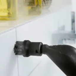 Czyszczenie fug parownicą z dyszą szczotkową, widoczna para, tło z kosmetykami i baterią umywalkową.