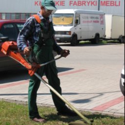 Agencja ochrony Skorpion sp. z o.o. - Mężczyzna w okularach i czapce przycina trawę podkaszarką spalinową na tle parkingu i budynku fabryki mebli.