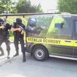 Dwóch umundurowanych ochroniarzy w pełnym rynsztunku, z bronią w rękach, pozuje obok firmowego samochodu z napisem 'Agencja Ochrony' i grafiką mapy świata w tle.