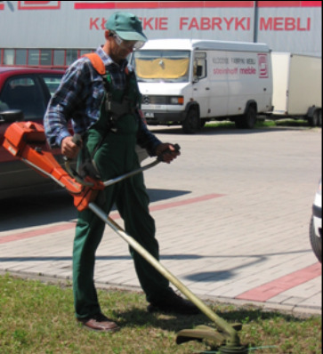 Mężczyzna w okularach i czapce przycina trawę podkaszarką spalinową na tle parkingu i budynku fabryki mebli.