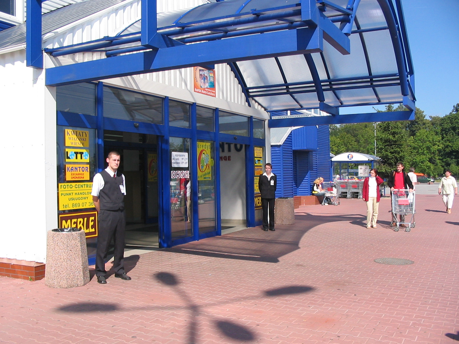 Dwóch ochroniarzy w czarnych uniformach stoi przed wejściem do budynku handlowego z niebieską konstrukcją nad drzwiami; w tle widać osoby z wózkami na tle zieleni.
