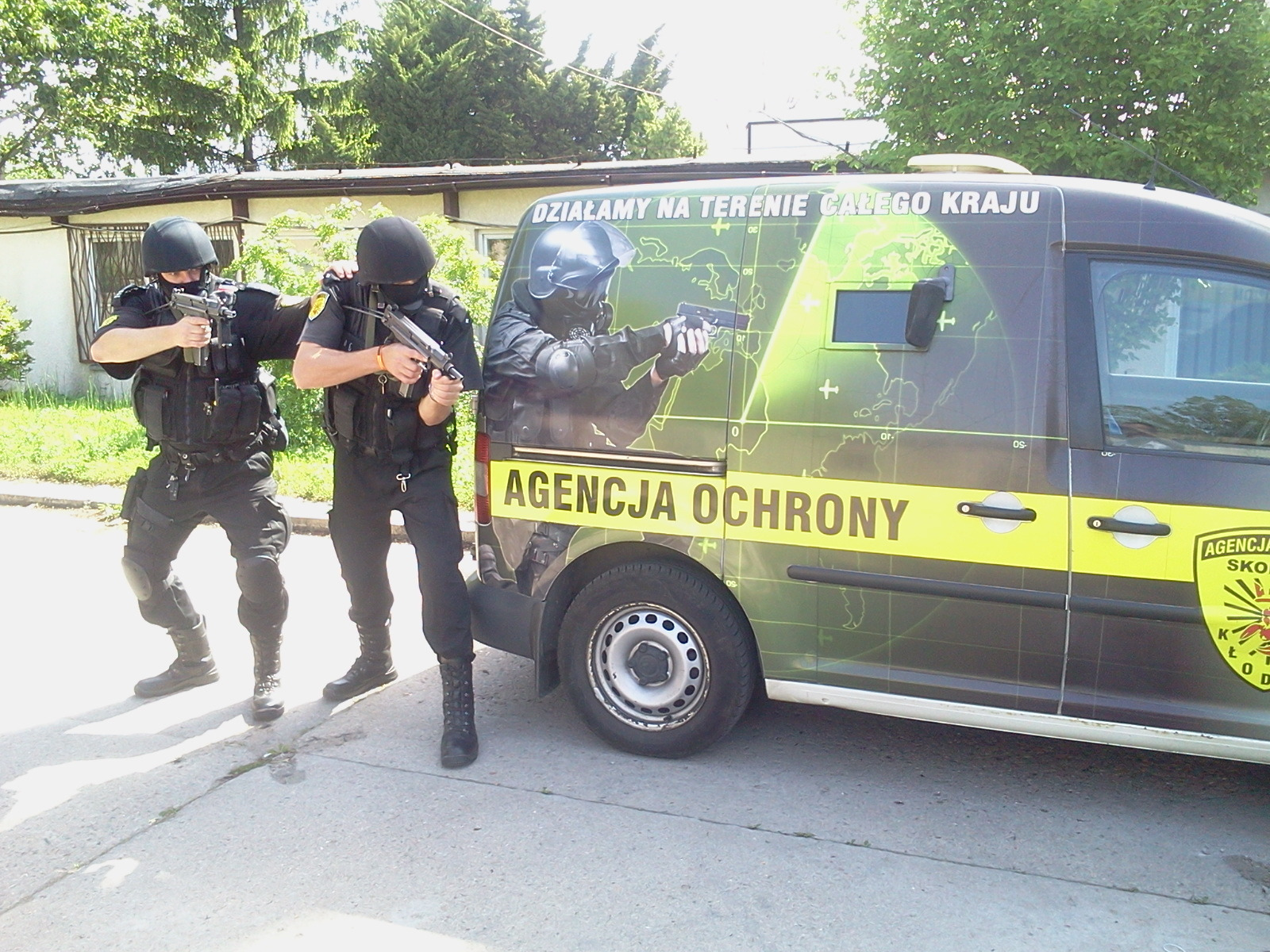 Dwóch umundurowanych ochroniarzy w pełnym rynsztunku, z bronią w rękach, pozuje obok firmowego samochodu z napisem 'Agencja Ochrony' i grafiką mapy świata w tle.