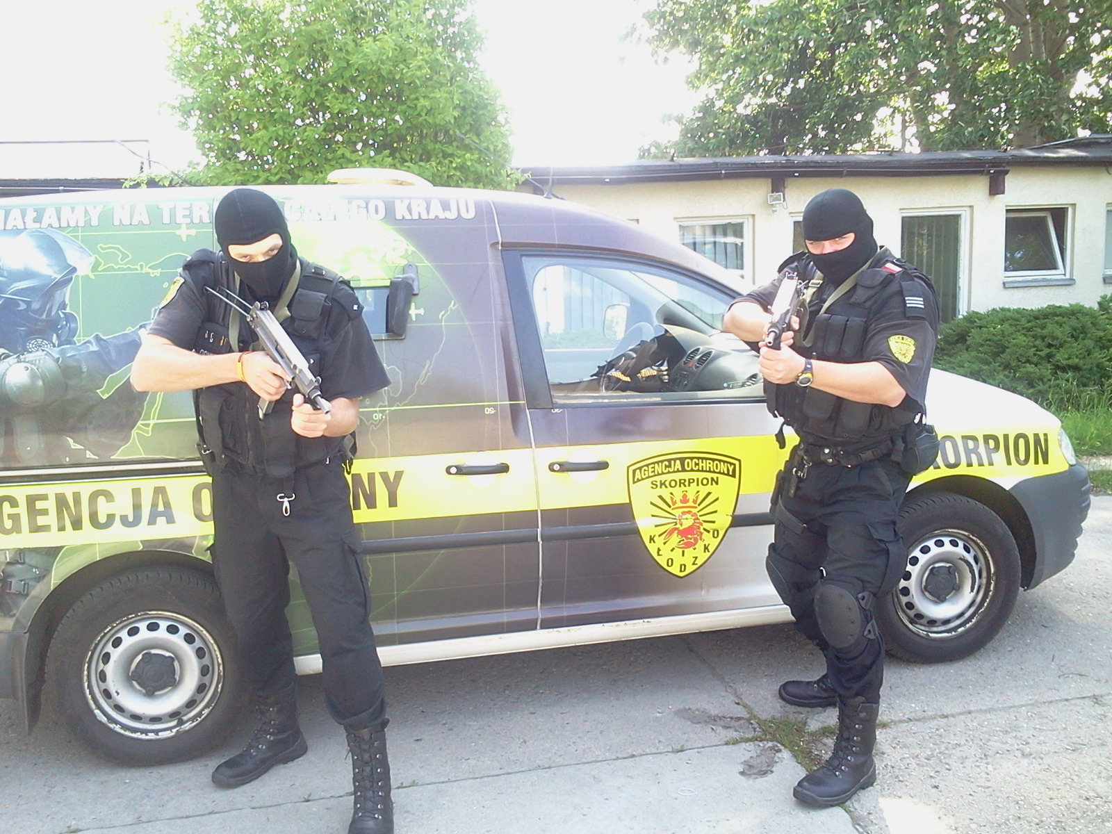 Dwóch umundurowanych ochroniarzy w kominiarkach, uzbrojonych w pistolety maszynowe, stoi przed samochodem Agencji Ochrony Skorpion z logo firmy na drzwiach.