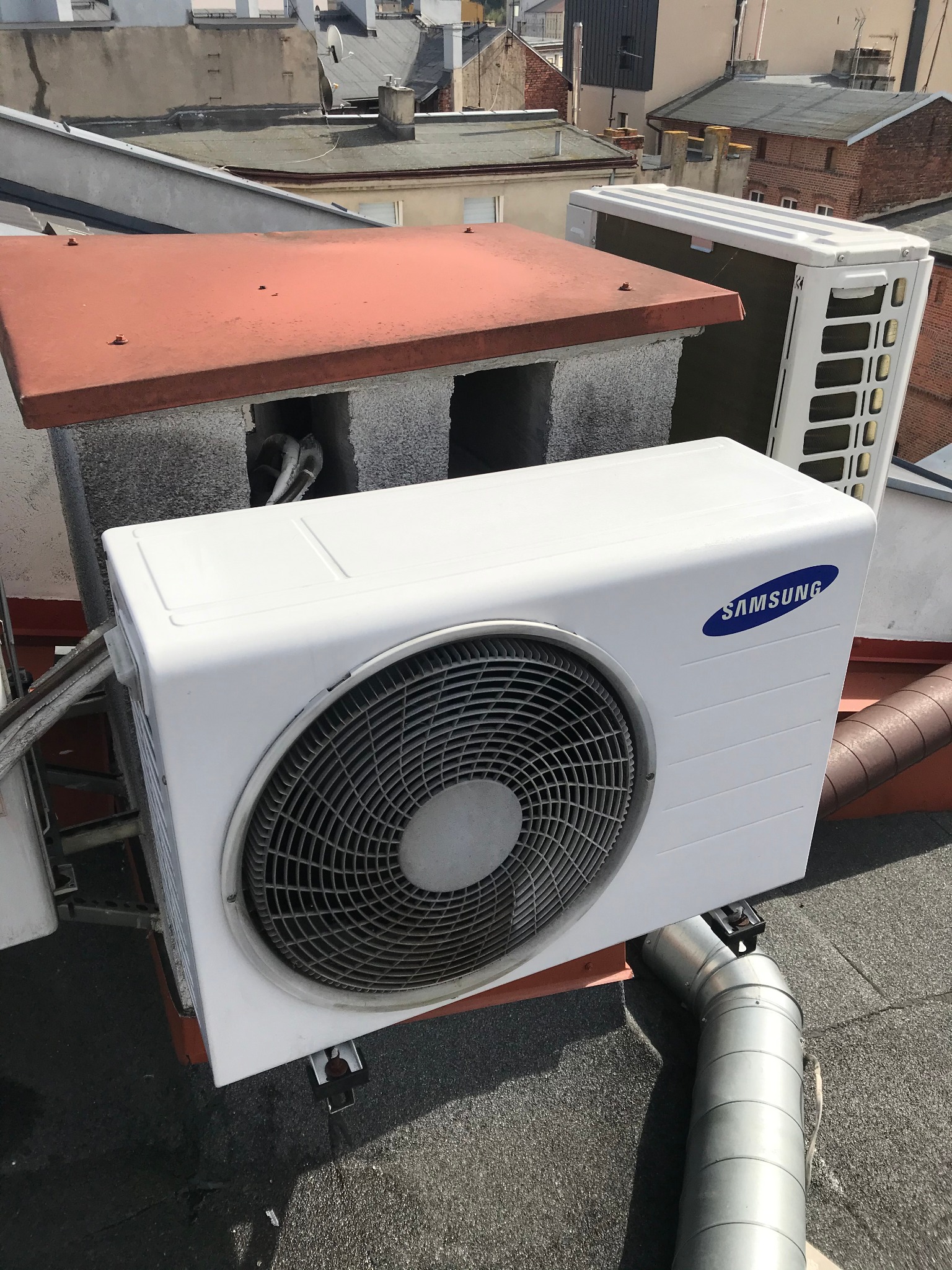 Biała jednostka zewnętrzna klimatyzacji Samsung na dachu budynku z widocznymi rurami i elementami montażowymi, na tle innych budynków.