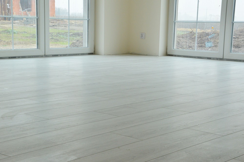 Jasne panele podłogowe ułożone w nowym pokoju z dużymi oknami, widok na zewnątrz z niedokończonym ogrodem, gniazdko elektryczne na ścianie.