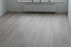 Jasnoszara podłoga z paneli ułożona w pustym, białym pokoju z grzejnikiem pod oknem.