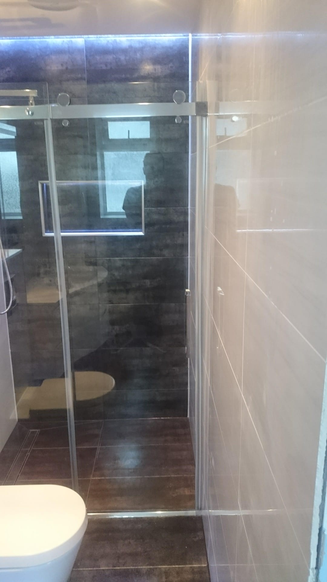 Nowoczesna kabina prysznicowa z przesuwnymi drzwiami ze szkła, widoczne ciemne płytki imitujące drewno na ścianie prysznica i podłodze, jasne płytki na pozostałej części ściany, podświetlenie LED...