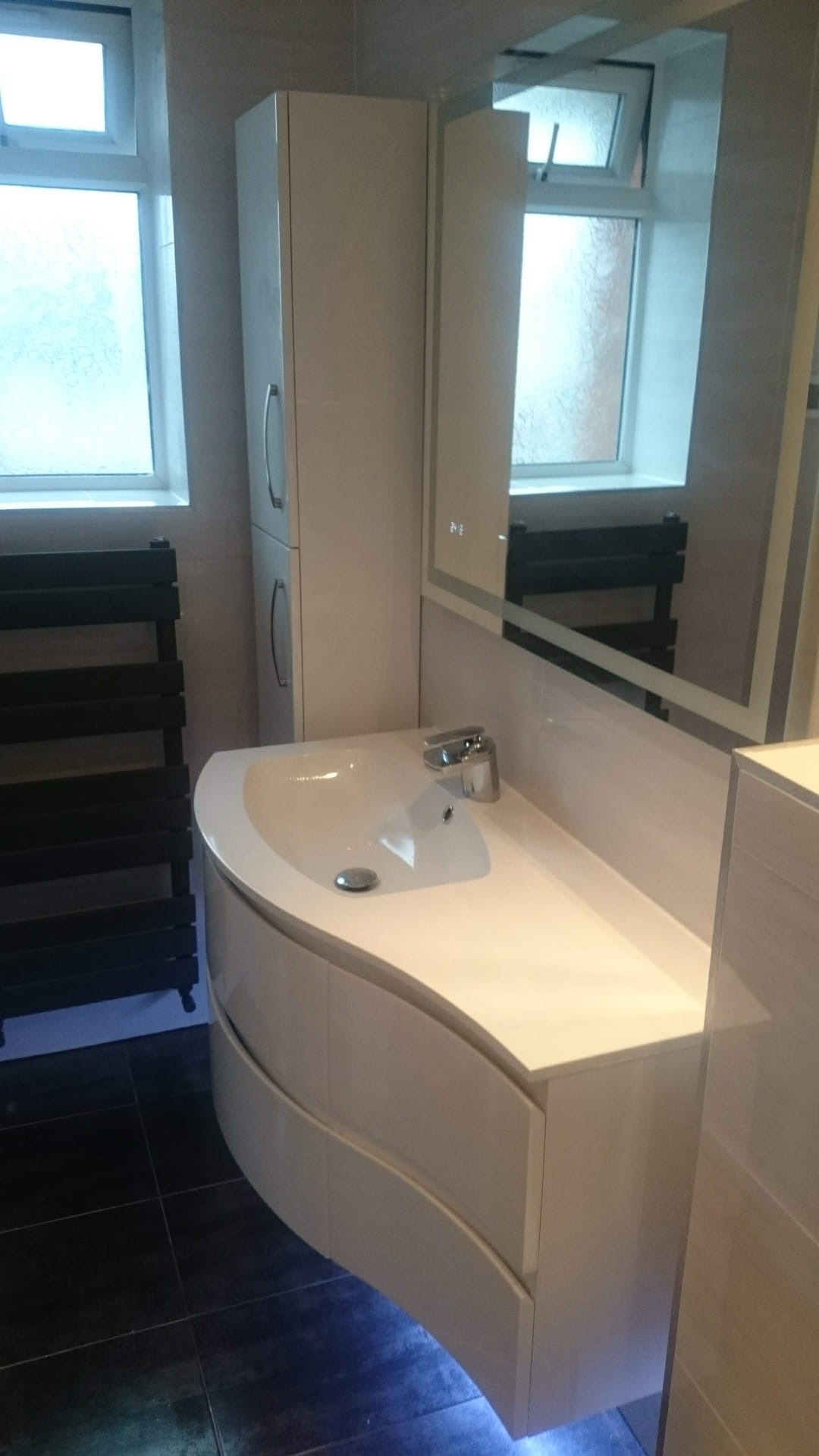 Biała, nowoczesna umywalka z szafką o zaokrąglonym kształcie, podświetlona od dołu niebieskim światłem, obok grzejnik drabinkowy i szafka wisząca, widoczne w lustrze odbicie grzejnika.