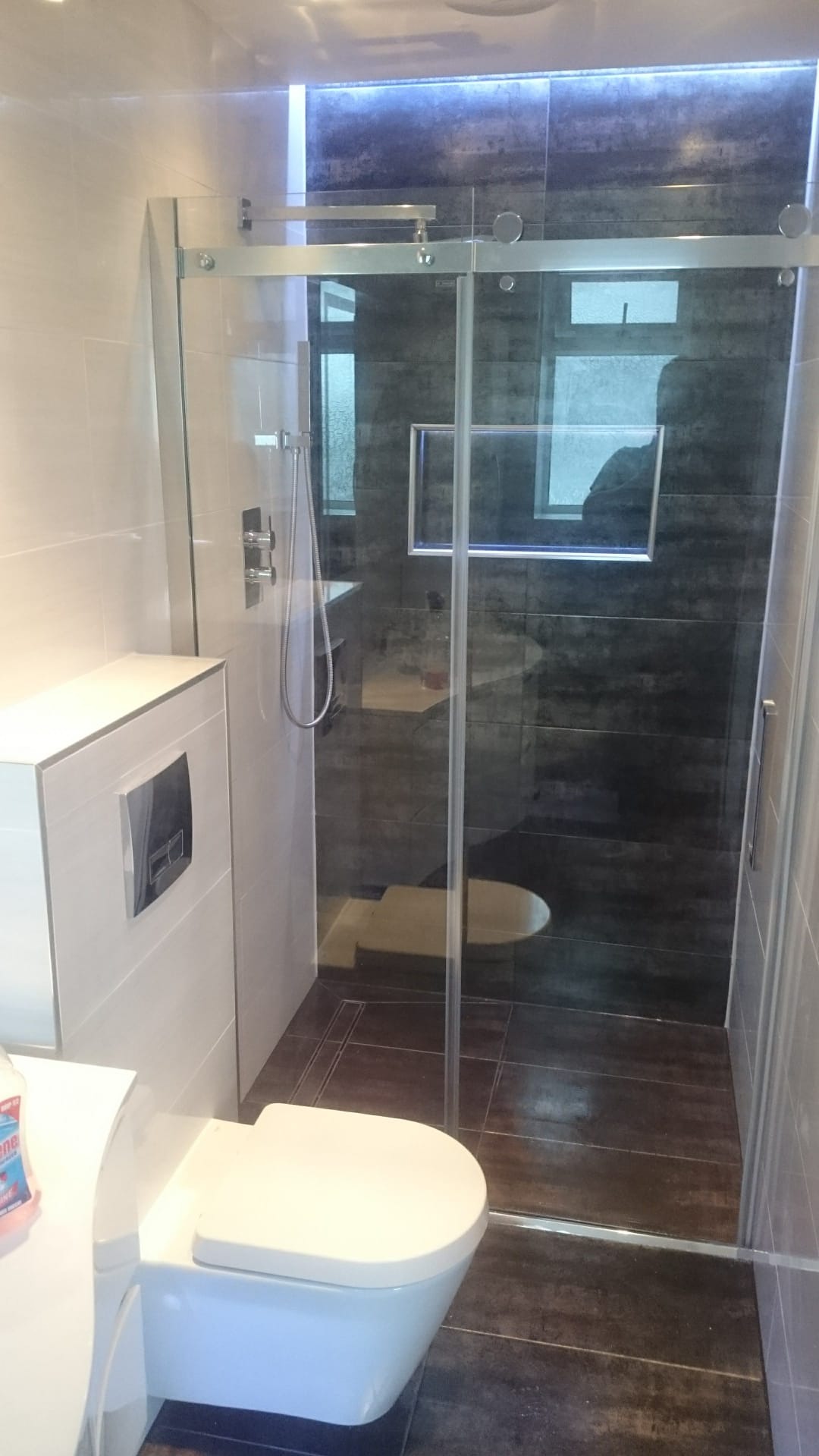 Nowoczesna łazienka z kabiną prysznicową ze szklanymi drzwiami przesuwnymi, toaleta podwieszana i ciemne płytki w strefie prysznica z podświetleniem LED.