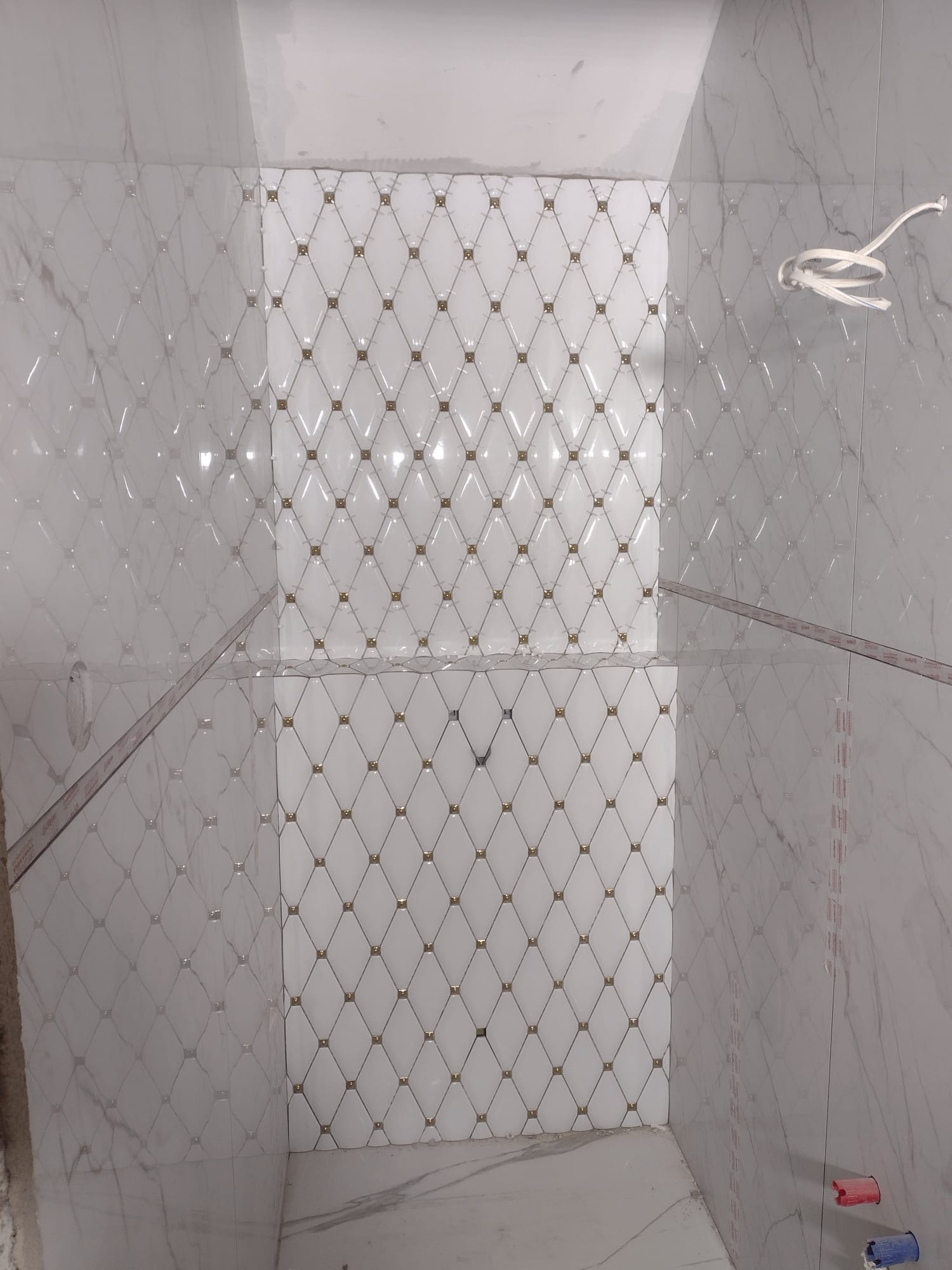 Wykończona ściana kabiny prysznicowej z białych płytek z motywem rombów i złotymi akcentami, widoczne przyłącza wodne i wystające przewody elektryczne.