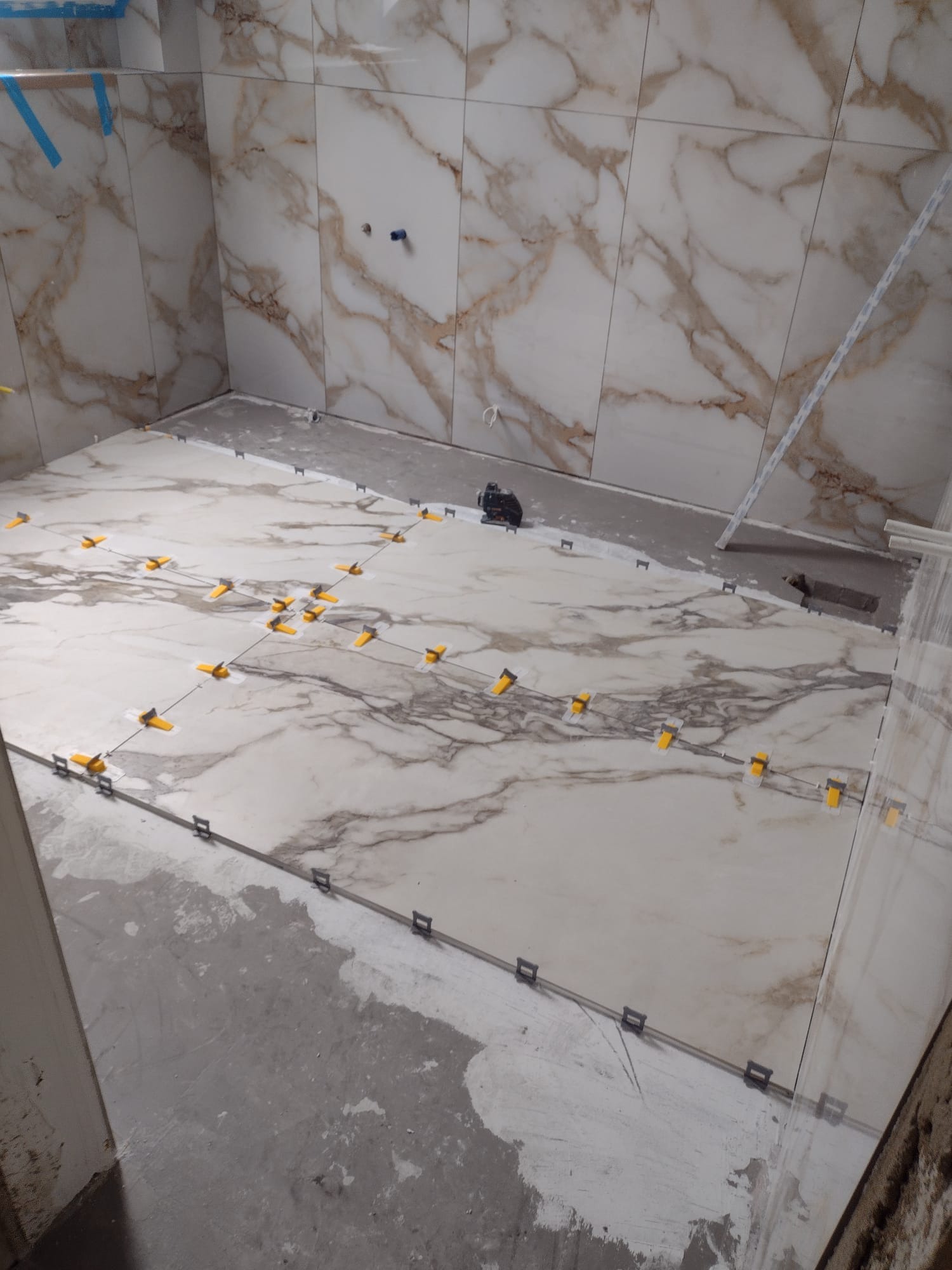 Układanie płytek imitujących marmur w łazience, widoczne żółte i czarne klipsy poziomujące, fragment ściany z wyprowadzonymi instalacjami hydraulicznymi.