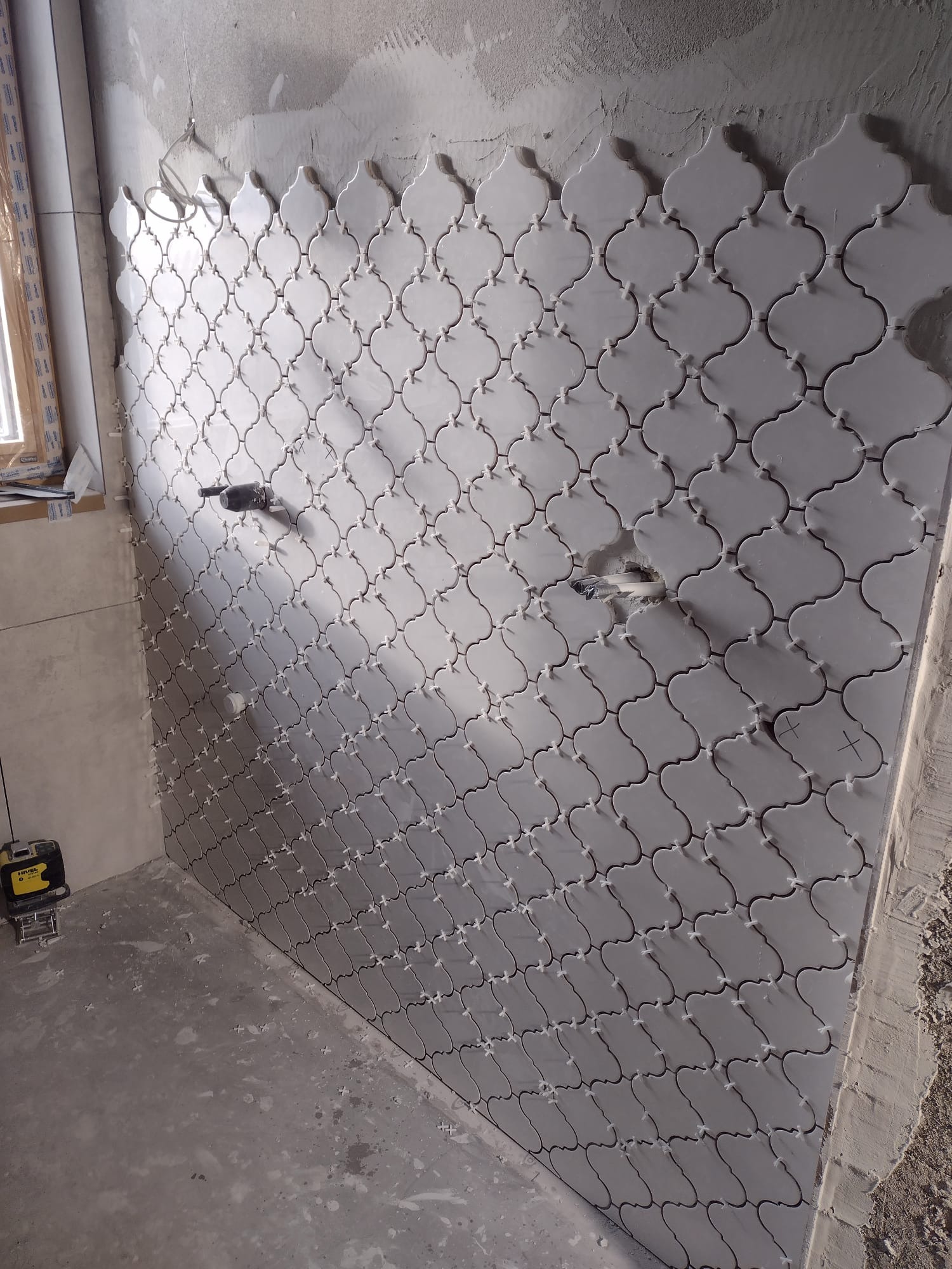 Ściana w trakcie układania dekoracyjnych, szarych płytek o marokańskim wzorze, z widocznymi krzyżykami dystansowymi i wystającymi przewodami elektrycznymi, na tle surowej, szarej ściany i podłogi...