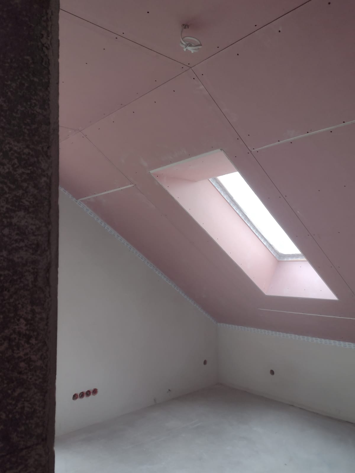 Poddasze w trakcie prac wykończeniowych: zamontowane płyty gipsowo-kartonowe na skosach dachu z wyciętym otworem na okno dachowe, widoczne łączenia płyt i przygotowane punkty elektryczne na suficie.