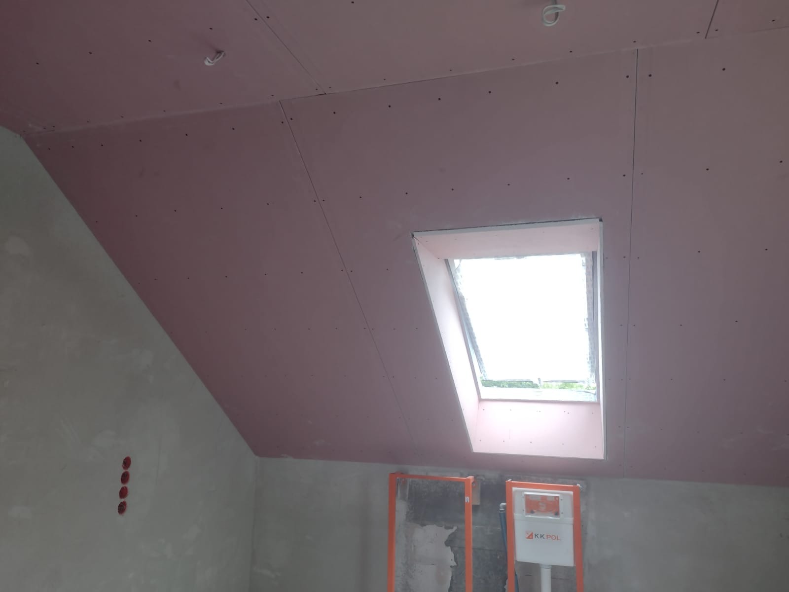 Poddasze z zamontowaną płytą karton-gips w kolorze różowym, widoczne okno dachowe i przygotowane miejsce pod instalację sanitarną.