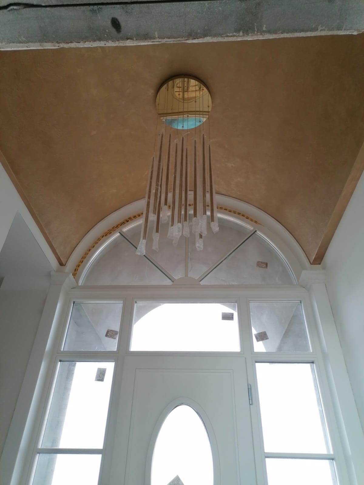 Wykończone wnętrze z gładzią na suficie w kolorze ochry, nowoczesnym żyrandolem z podłużnymi elementami i białymi drzwiami z łukowym oknem nad nimi.