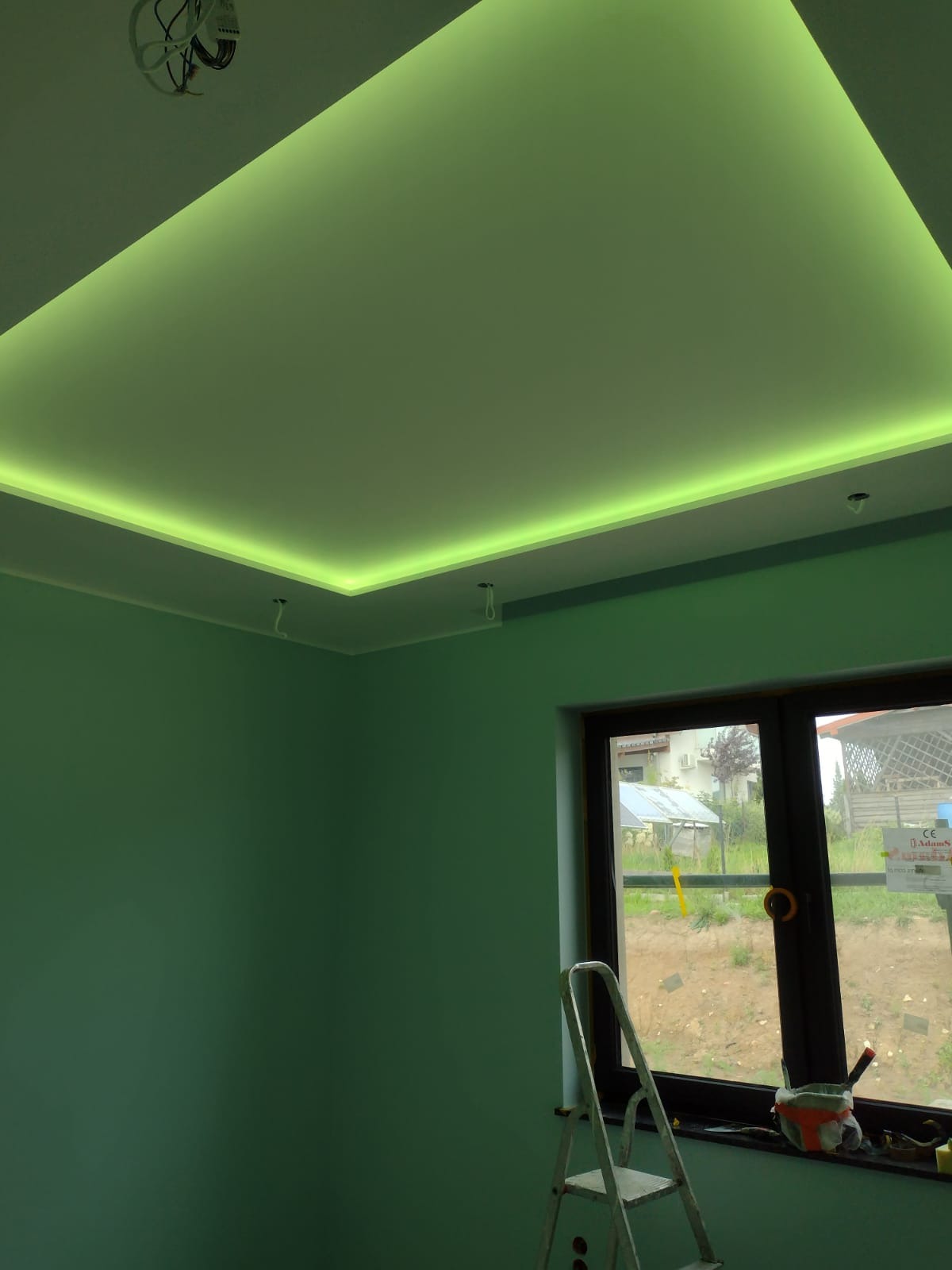 Pokój z pomalowanymi na zielono ścianami i sufitem podwieszanym z oświetleniem LED w kolorze zielonym, drabina oparta o ścianę przy oknie.