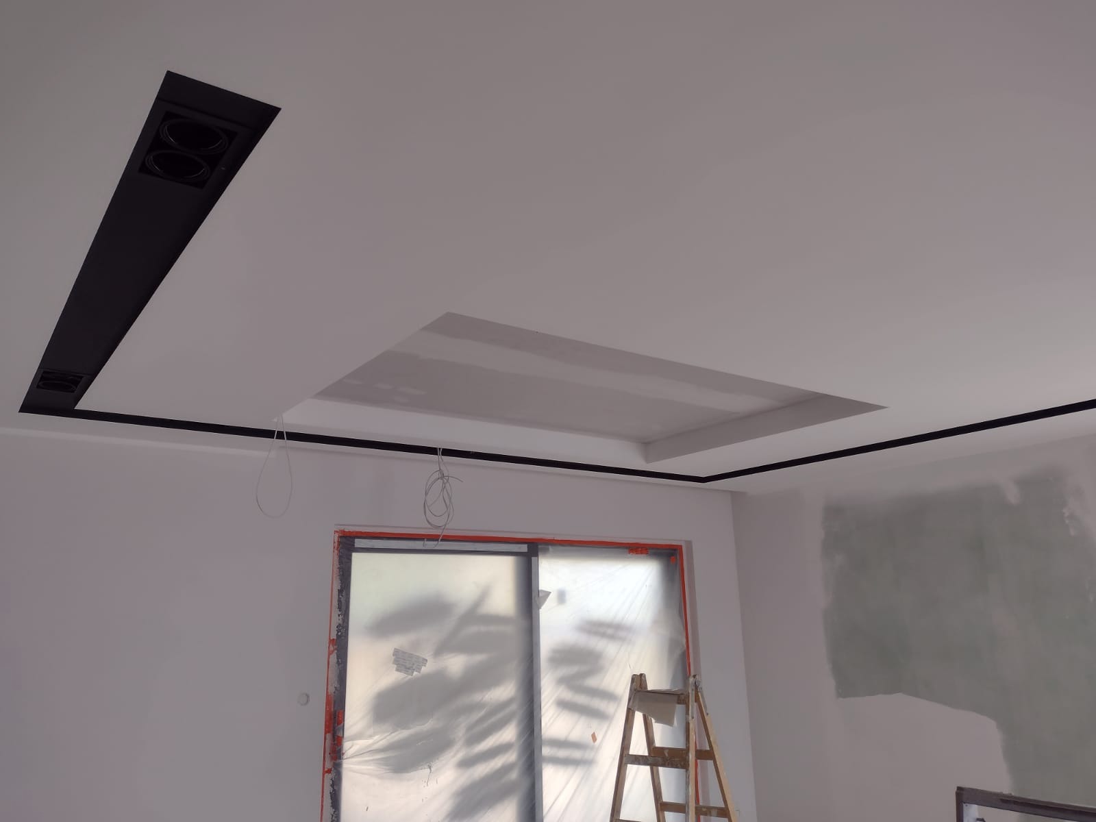 Wnętrze w trakcie prac wykończeniowych: sufit z podwieszanymi elementami i czarnymi listwami dekoracyjnymi, okno zabezpieczone folią, drabina drewniana w pomieszczeniu.