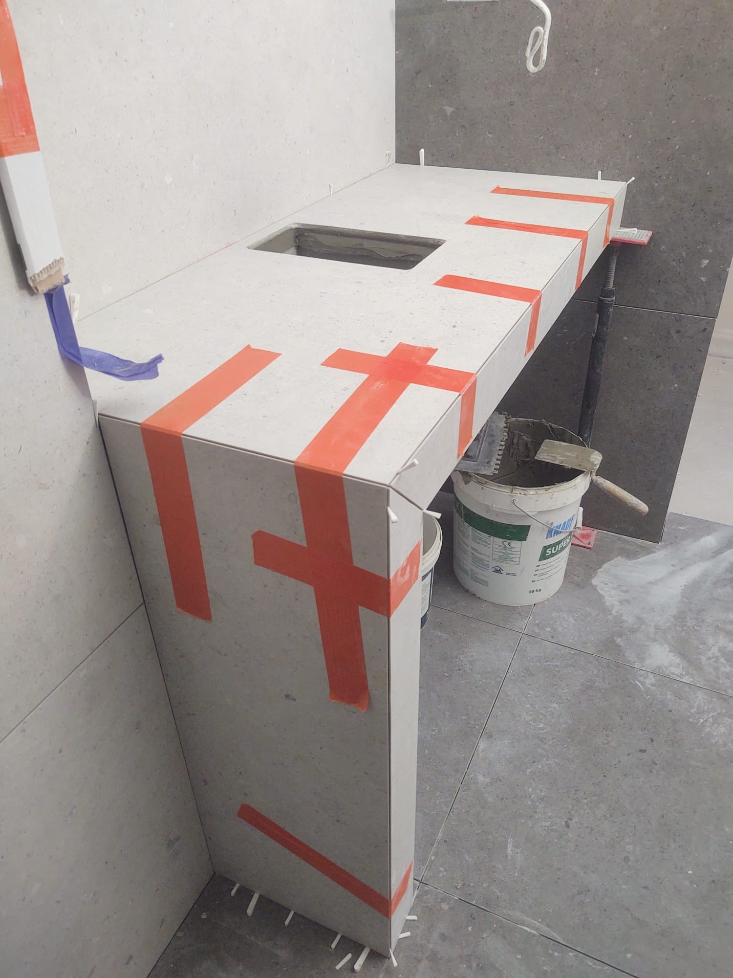 Montaż blatu łazienkowego z otworem na umywalkę, zabezpieczonego pomarańczową taśmą, z widocznym wiadrem z zaprawą i narzędziami w trakcie prac wykończeniowych.