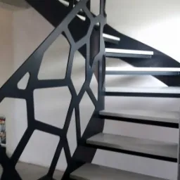 Spiralne schody z ażurową, metalową balustradą o geometrycznym wzorze, szare stopnie, białe ściany w tle.
