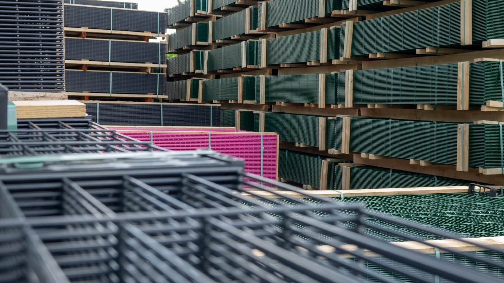 Stosy paneli ogrodzeniowych w różnych kolorach (czarny, zielony, różowy) ułożone na drewnianych paletach, widoczne elementy konstrukcyjne ogrodzeń na pierwszym planie.