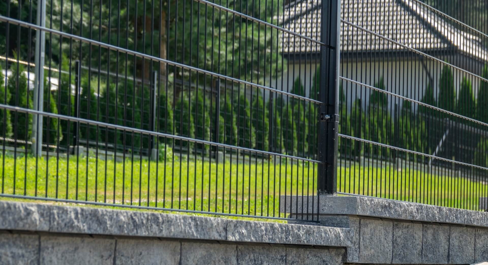 Czarny panelowy płot metalowy na betonowym murku, z widokiem na trawnik i tuje w tle.