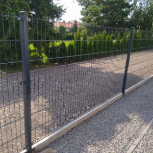 Szare panele ogrodzeniowe na betonowej podmurówce, widok z perspektywy gruntu, z tłem zielonych tuj i czerwonego dachu domu.