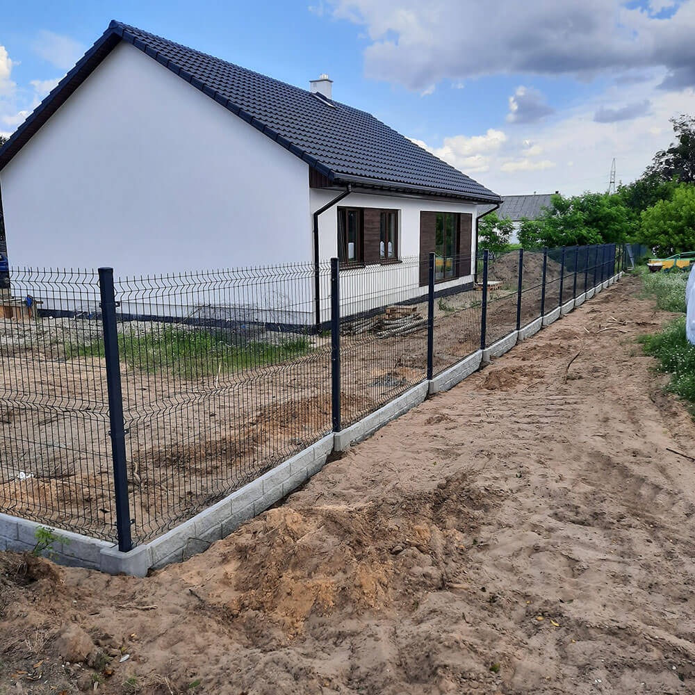 Nowoczesne ogrodzenie panelowe z podmurówką betonową otaczające posesję z białym domem i ciemnym dachem w słoneczny dzień.
