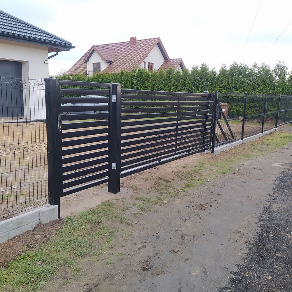 Nowoczesne, czarne ogrodzenie z poziomych profili metalowych i siatki, z bramą wjazdową, na tle domu z czerwoną dachówką i zielonych drzew.