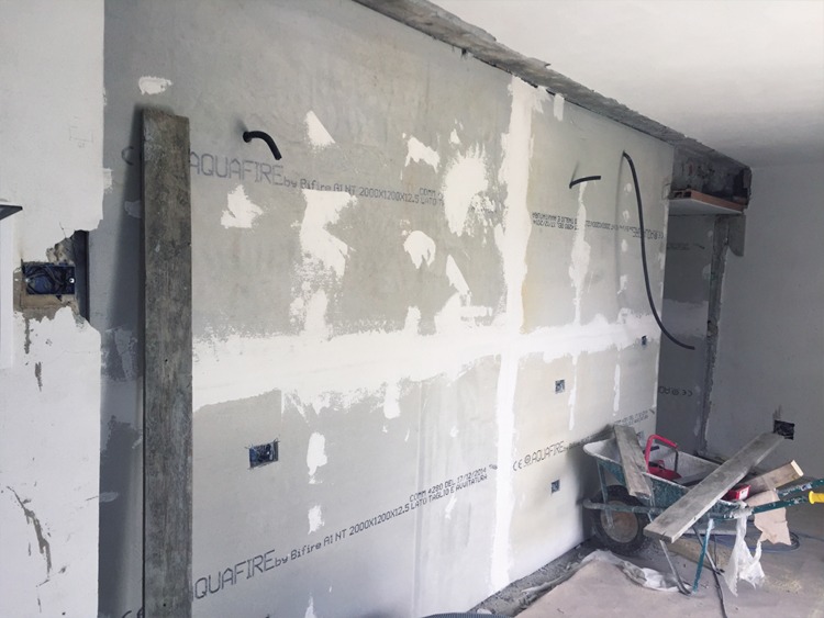 Wnętrze w trakcie remontu: ściany pokryte płytami AquaFire, widoczne przewody elektryczne, otwory na gniazdka, taczka z materiałami budowlanymi w rogu pomieszczenia.