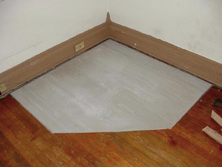 Fragment podłogi z odnowioną, pomalowaną na szaro częścią parkietu w rogu pokoju, widoczne listwy przypodłogowe i gniazdko elektryczne na ścianie.