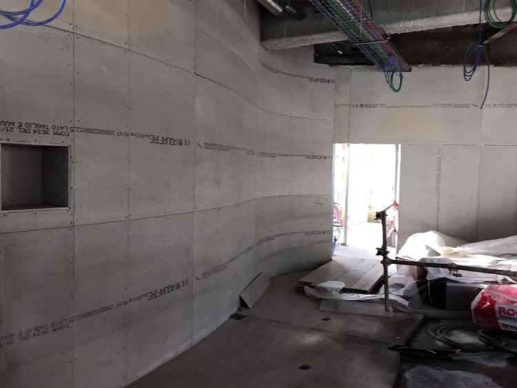 Wnętrze w trakcie wykańczania z zakrzywioną ścianą z płyt gipsowo-kartonowych, widoczne kable pod sufitem, otwór w ścianie i materiały budowlane na podłodze.