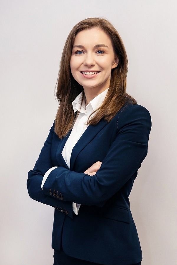 Portret uśmiechniętej kobiety w granatowym garniturze i białej koszuli, z rękami skrzyżowanymi na piersiach na jasnym tle.