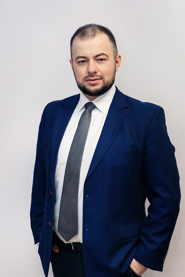 Portret mężczyzny w granatowym garniturze i wzorzystym krawacie na jasnym tle, z ręką w kieszeni.